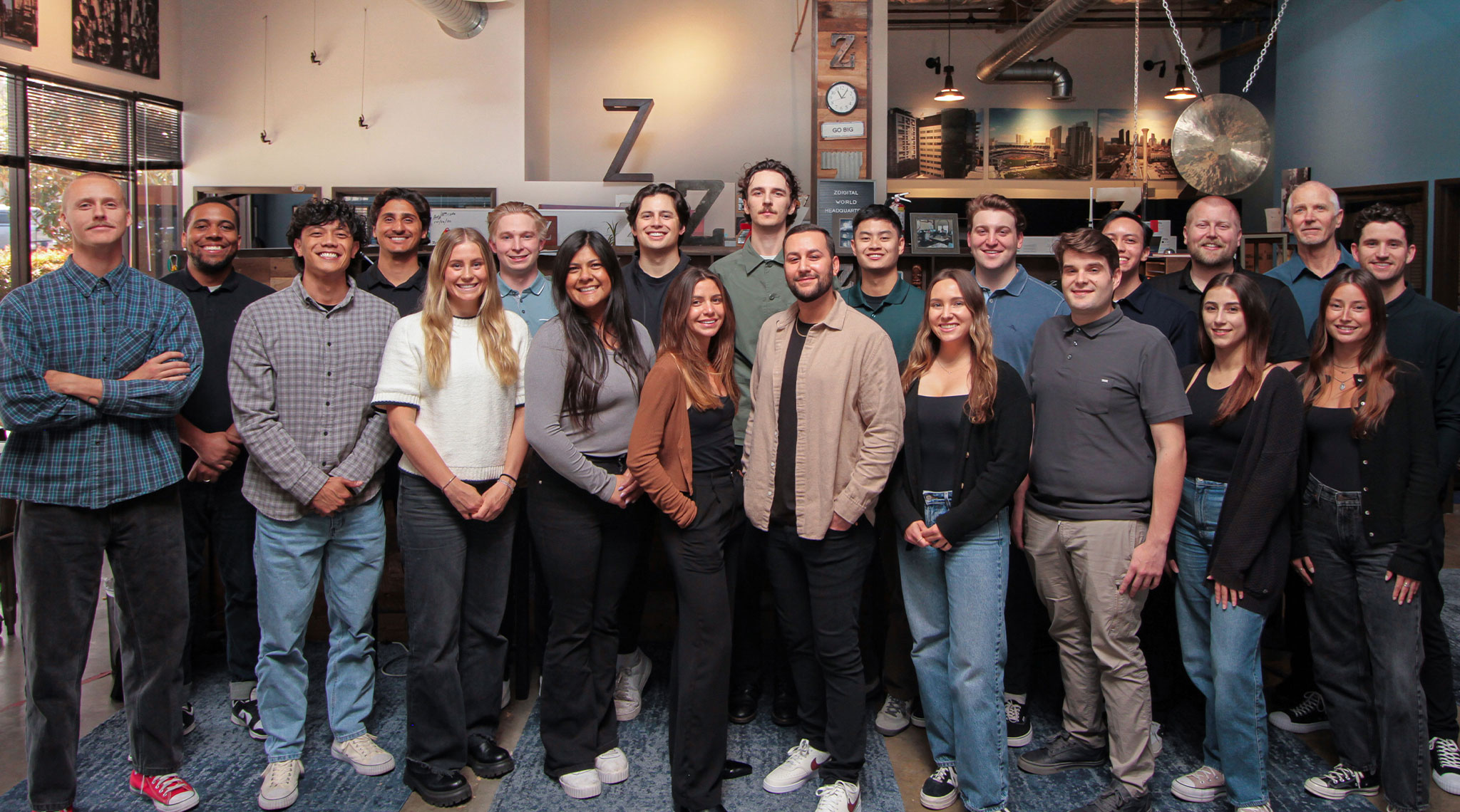 A diverse group of people standing indoors, posing for a group photo in casual attire. Some are smiling, and a large gong is visible in the background.